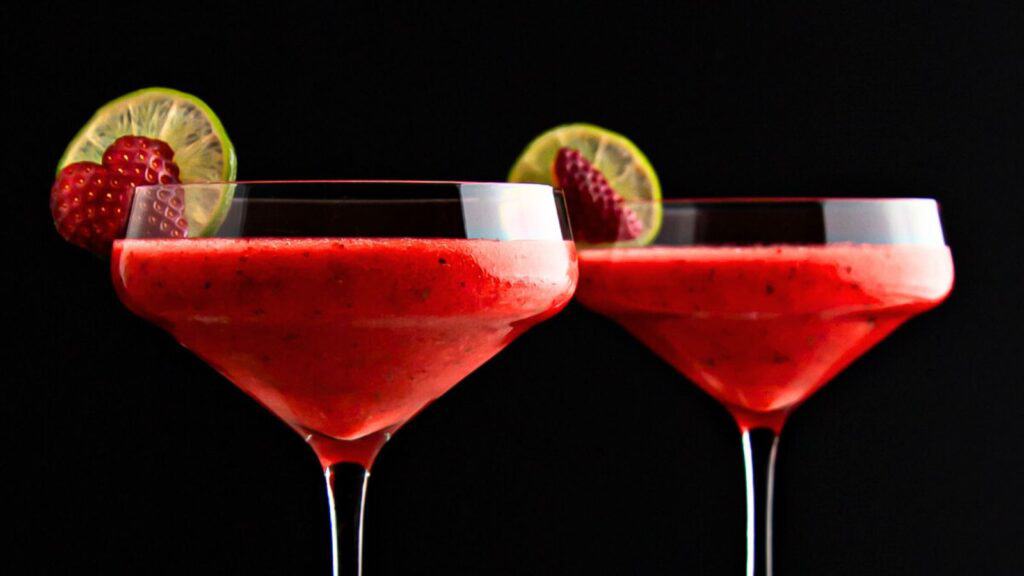 Two strawberry cocktails in martini glasses garnished with lime slices and raspberries, set against a black background.