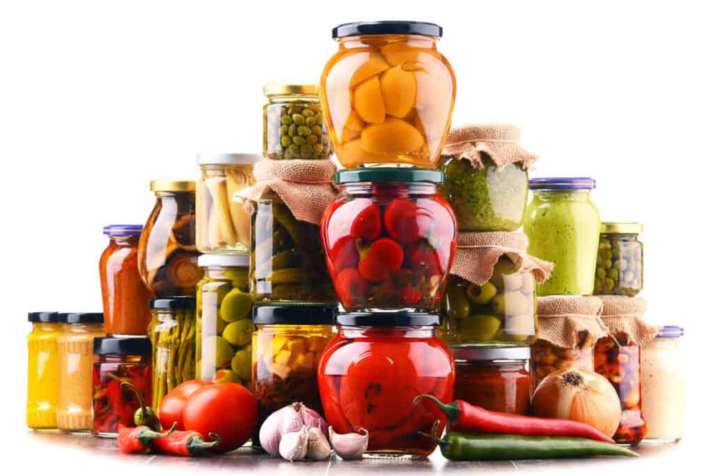 Assorted preserved vegetables in glass jars, including tomatoes, peppers, and pickles, isolated on a white background.