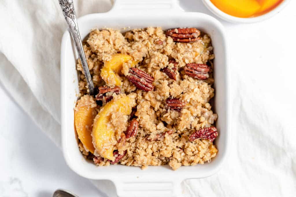 A square white dish filled with peach crumble topped with pecans and a spoon on a white cloth surface.