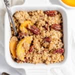 A square white dish filled with peach crumble topped with pecans and a spoon on a white cloth surface.