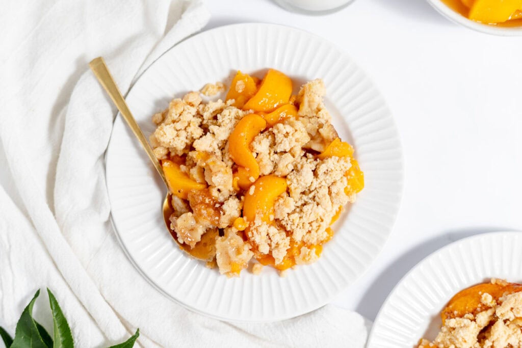 A white plate with a serving of peach cobbler, featuring golden peaches and a crumbly topping. A gold fork is placed on the side of the plate, which is set on a white cloth.