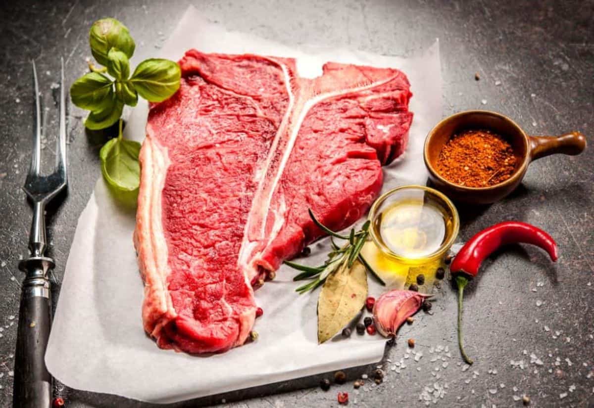 Raw t-bone steak on parchment paper, surrounded by a fork, basil leaves, mixed spices, oil, garlic, bay leaves, rosemary, and a red chili pepper.