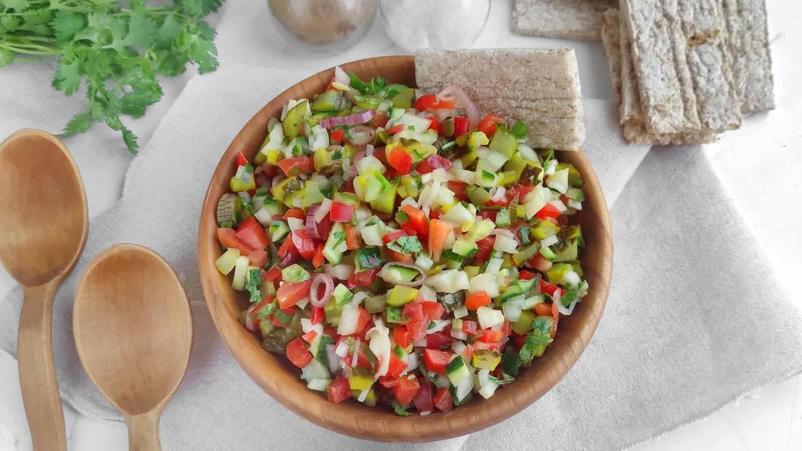 A wooden bowl filled with pickle de gallo salsa, surrounded by whole grain crackers and wooden utensils, on a white cloth.