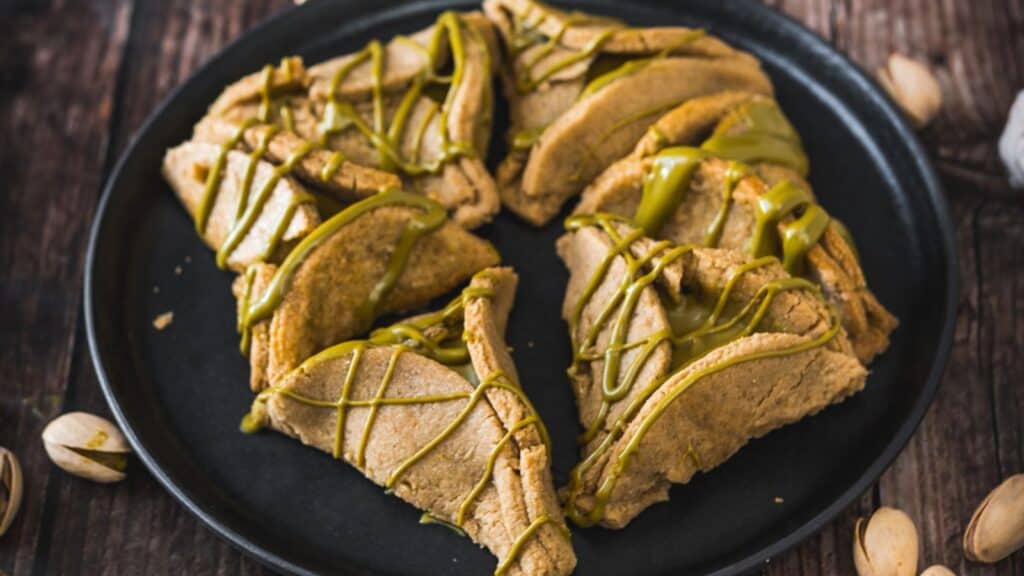Slices of pistachio cake with a drizzle of green syrup, served on a round black plate, accompanied by whole pistachios.