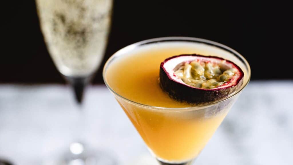 A yellow cocktail in a martini glass is garnished with a half passion fruit. A flute of sparkling wine is in the background.