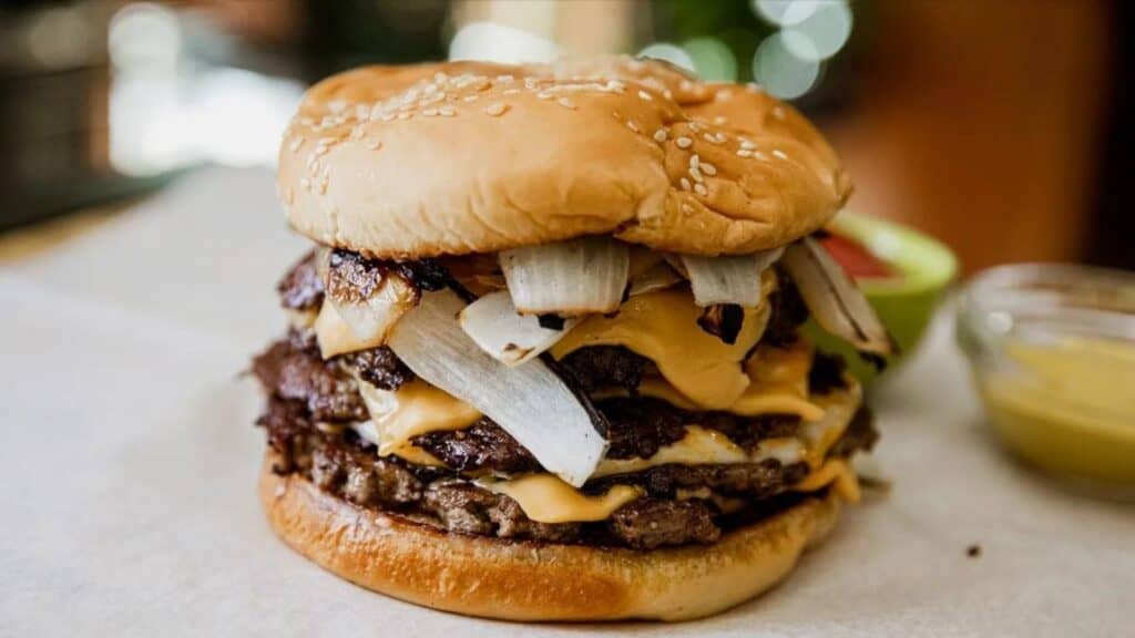 A Quadruple Smash Burger with melted cheese, grilled onions, and a sesame seed bun is placed on a white surface. Condiments in the background are slightly blurred.