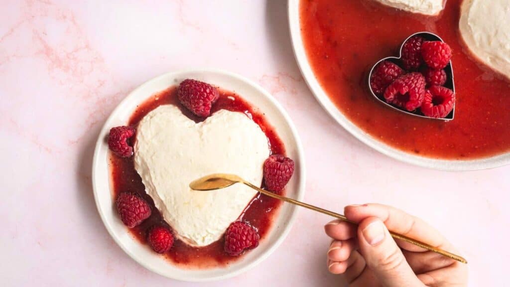 A heart-shaped cheesecake topped with raspberries on a red sauce, with a hand holding a spoon on a pink background.