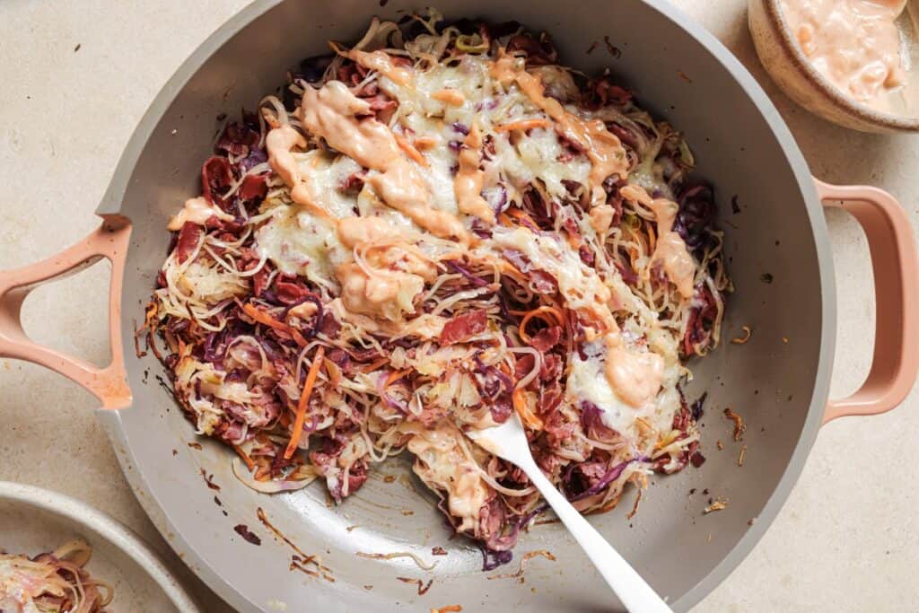 Sautéed red and white cabbage in a pan, topped with melted cheese and a drizzle of pink sauce, with a serving spoon.