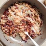 Sautéed red and white cabbage in a pan, topped with melted cheese and a drizzle of pink sauce, with a serving spoon.
