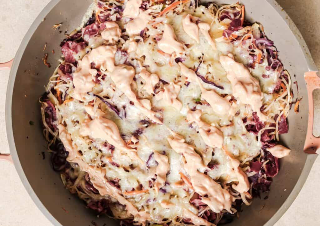 Cooked cabbage and carrot slaw in a skillet, topped with melted cheese and drizzled with pink sauce.