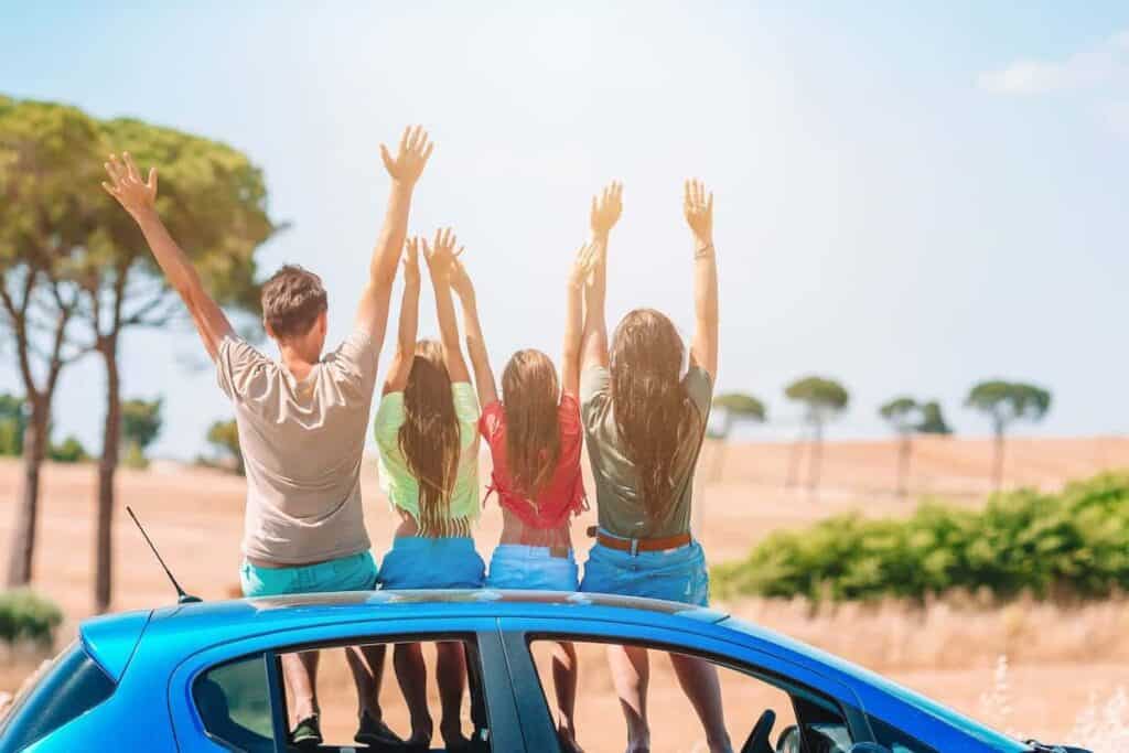 A family of four raising their hands joyfully while sitting on a blue car against a scenic, sunny countryside backdrop.