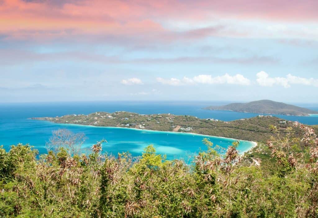 A scenic view of a hilly coastal landscape with turquoise waters, islands in the distance, and a vivid sky featuring shades of orange and blue. Greenery is visible in the foreground.