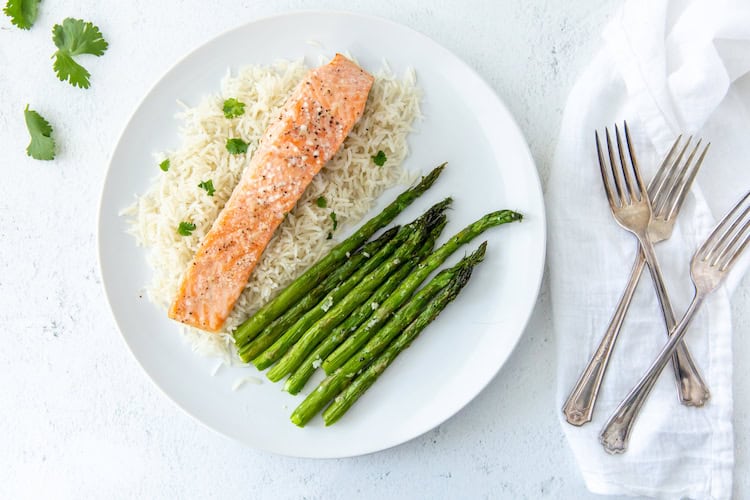 A plate with grilled salmon, asparagus, and rice, garnished with parsley, on a white table with two forks beside it.