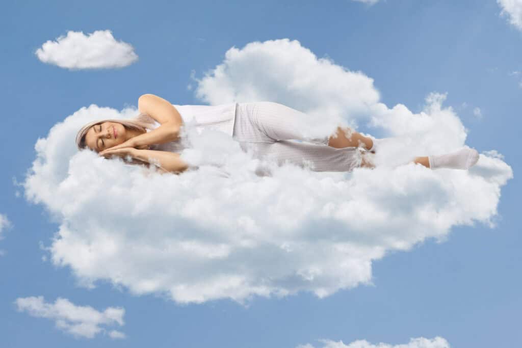A woman in pajamas is lying down and sleeping on a cloud against a blue sky.