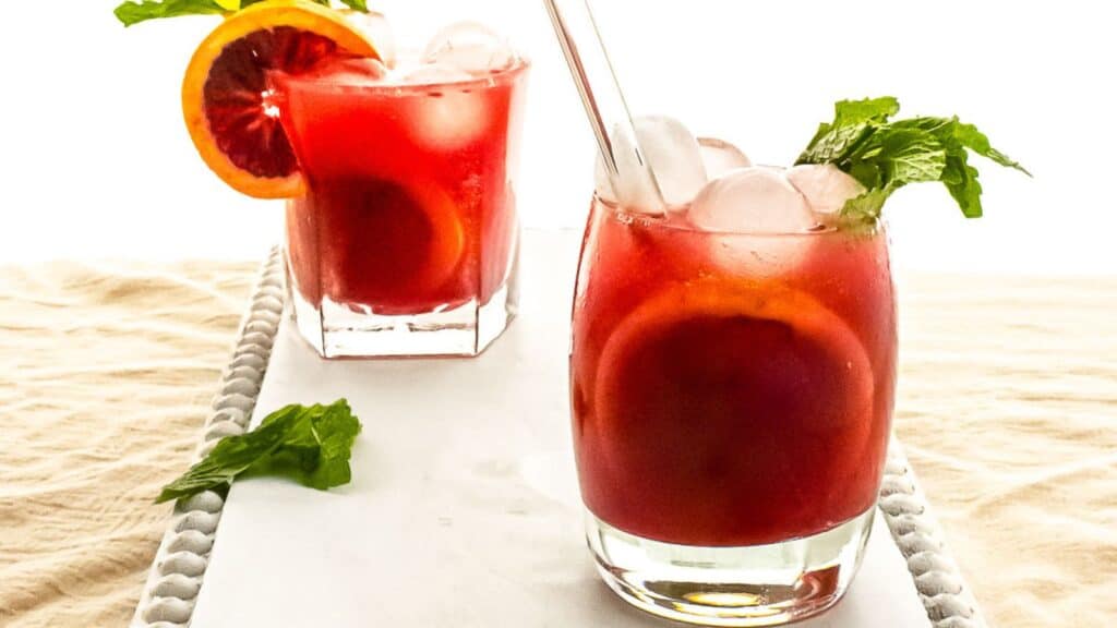 Two glasses of iced red fruit punch garnished with mint leaves and a lemon slice, placed on a white surface with a woven mat underneath.
