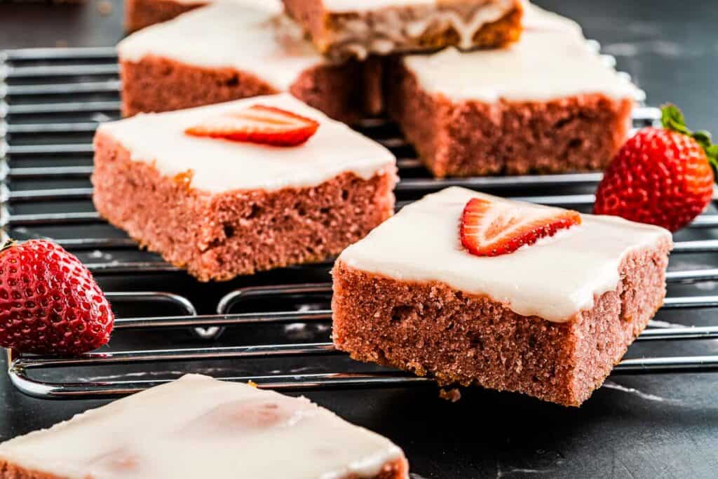 Sliced strawberry-topped brownies with white icing on a cooling rack, with fresh strawberries nearby.
