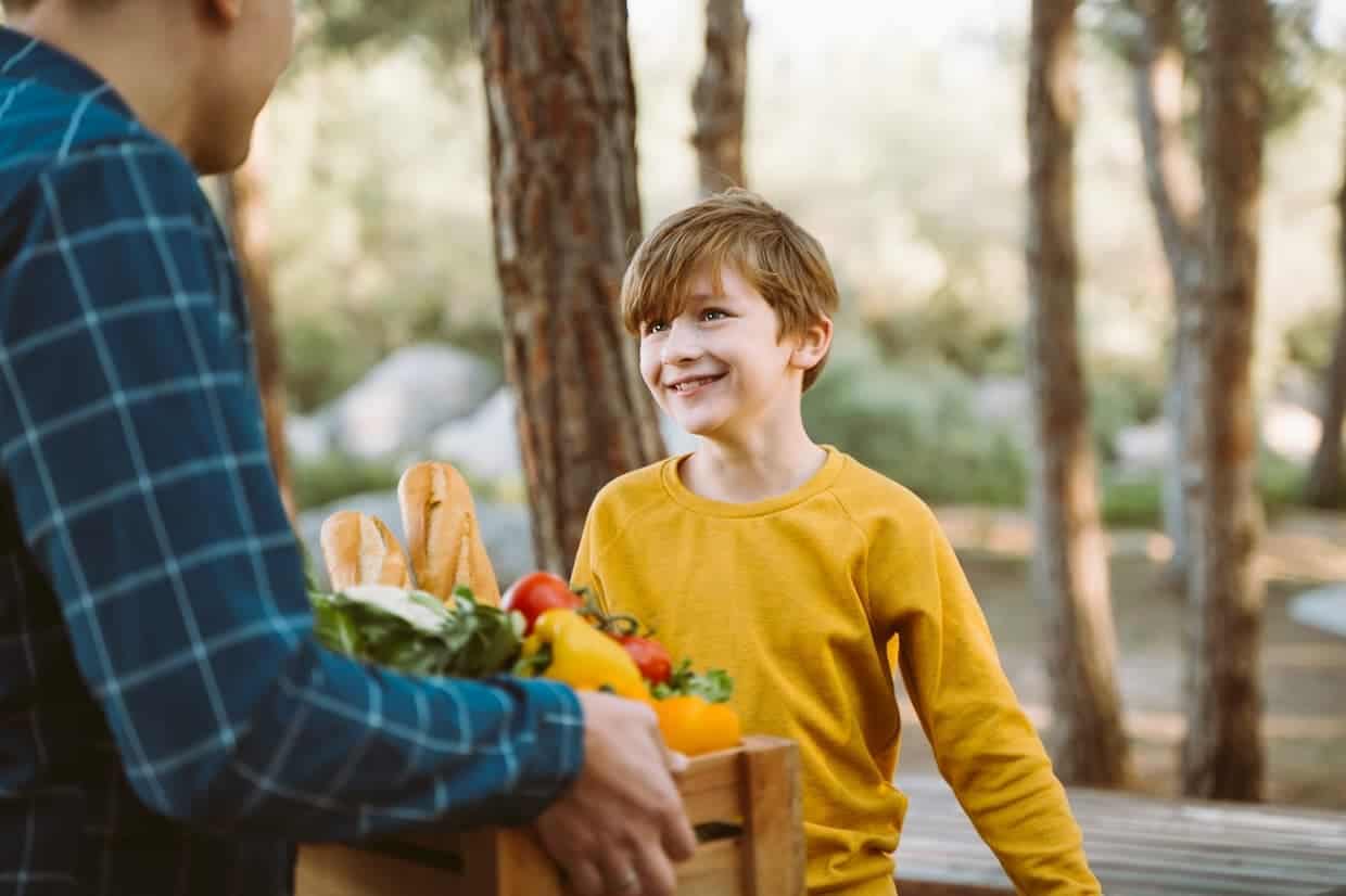 A child in a yellow shirt smiles while a person in a plaid shirt holds a crate of groceries with vegetables and baguettes in an outdoor setting.