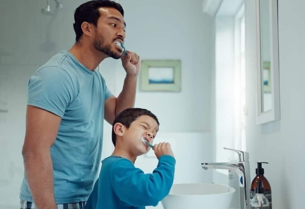 A man and a young boy are brushing their teeth together in a bathroom with a closed faucet.