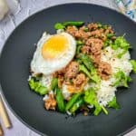 A plate of Thai pork & Green beans with rice, topped with a fried egg, served with chopsticks on the side.