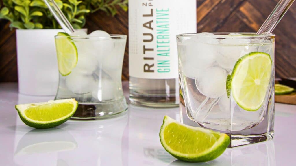 Two glasses of clear liquid with lime slices and ice, a bottle labeled "ritual gin alternative", and lime wedges on a wooden table.