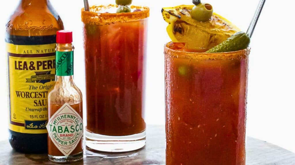 Two bloody mary cocktails with garnishes, next to bottles of worcestershire and tabasco sauce.