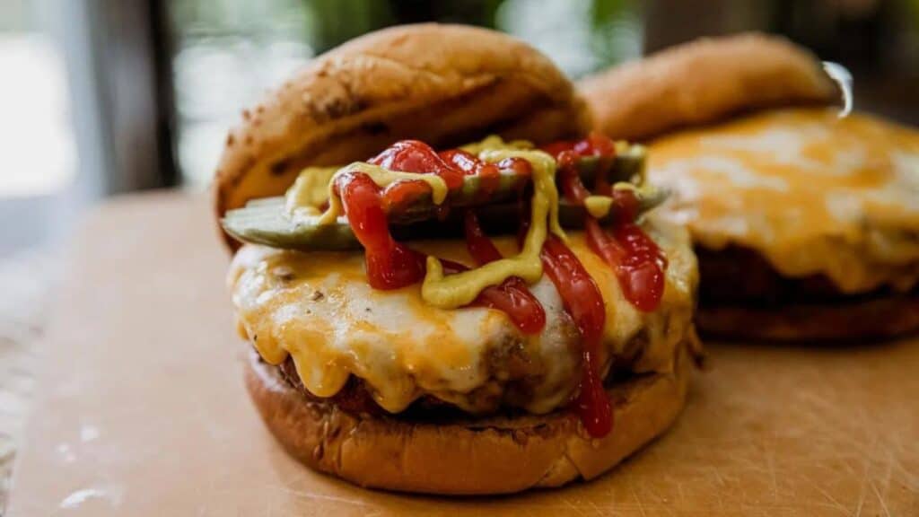 Close-up of a cheeseburger topped with ketchup, mustard, and pickles on a wooden surface. Another similar cheeseburger is in the background.