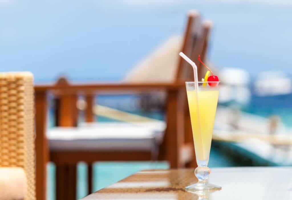 A tall glass of yellow tropical drink with a cherry and straw on a table, with blurred outdoor seating and ocean in the background.