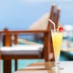A tall glass of yellow tropical drink with a cherry and straw on a table, with blurred outdoor seating and ocean in the background.