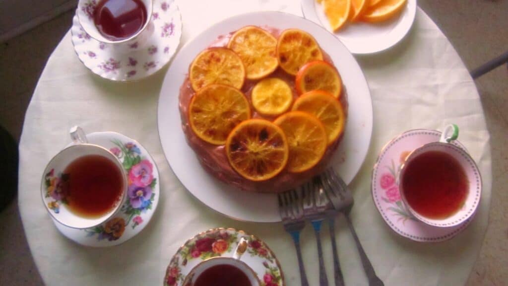 A round table with cups of tea, slices of orange, and an orange-topped cake along with three forks on plates.