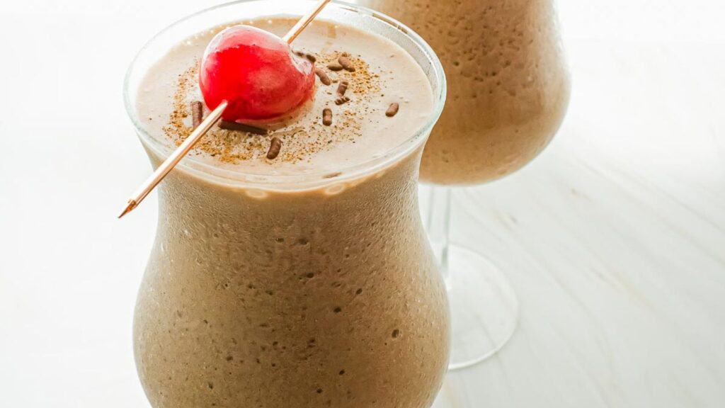 Two glasses of chocolate milkshake garnished with a cherry on top, served on a white surface.