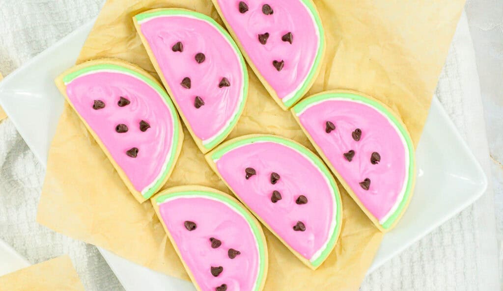 Decorated summertime cookies shaped like watermelon slices with pink icing and black sprinkle seeds on a white plate.