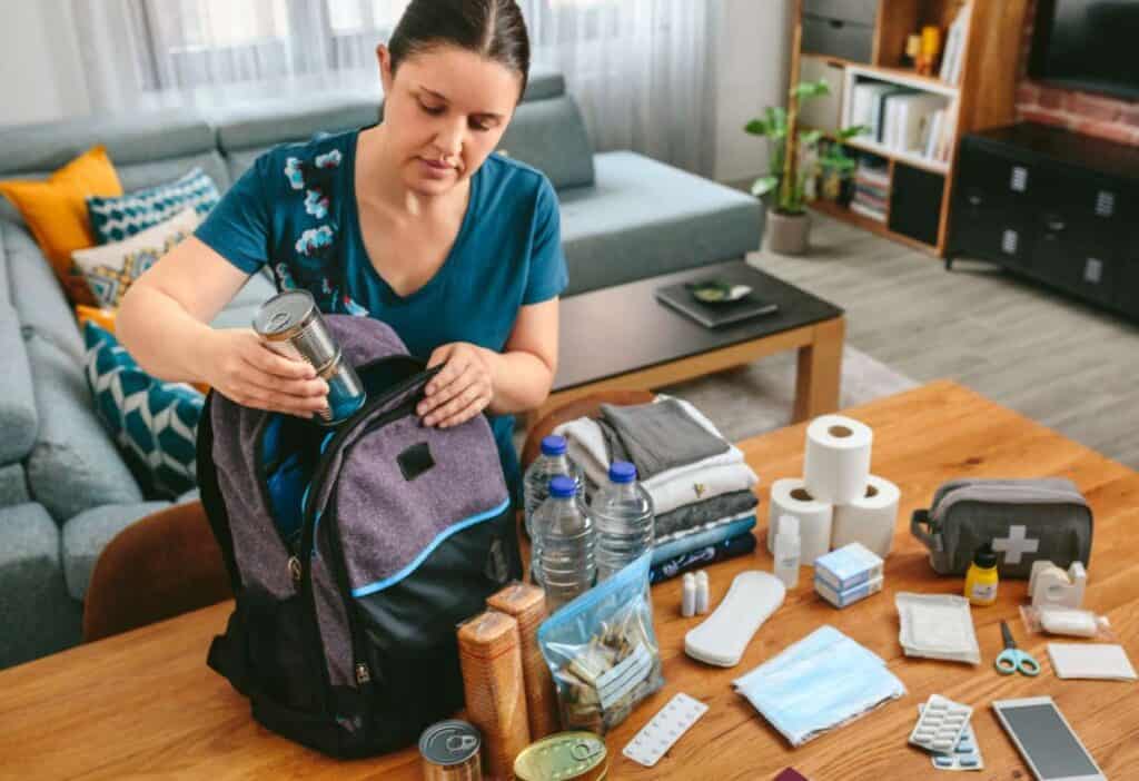A woman packs a gray and black backpack with canned food on a wooden table filled with survival items including water bottles, toiletries, and medical supplies inside a living room.