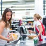 A woman in a white top checks out groceries with a cashier in a red apron at a supermarket.