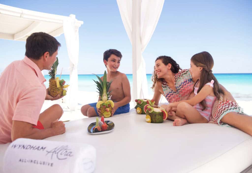 A family of four relaxes on a beach cabana, enjoying tropical drinks served in pineapple and coconut shells, with the ocean in the background.