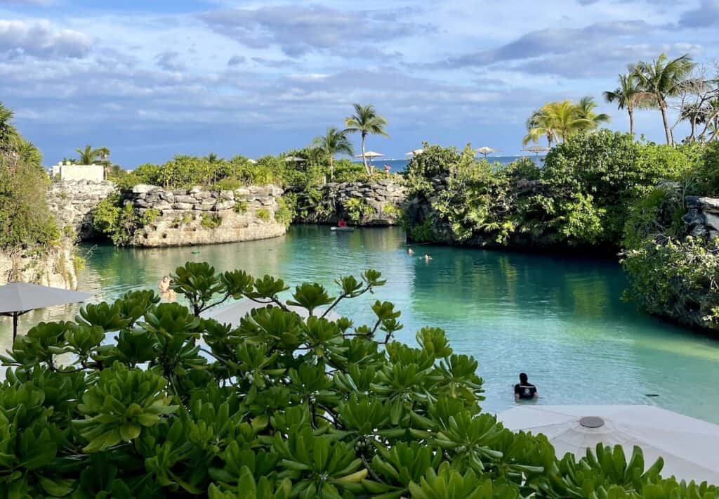 A serene lagoon surrounded by lush greenery with a few people enjoying the water, under a partly cloudy sky. Palms and rocky cliffs line the edges.
