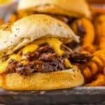 A beef sandwich with melted cheese on a bun, served with curly fries on a metal tray.