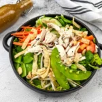 A bowl of Chinese chicken salad with shredded chicken, snap peas, carrots, and peppers, accompanied by a bottle of dressing on the side.