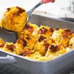 A hand scooping a serving of baked cheesy bread rolls topped with bacon from a rectangular dish.