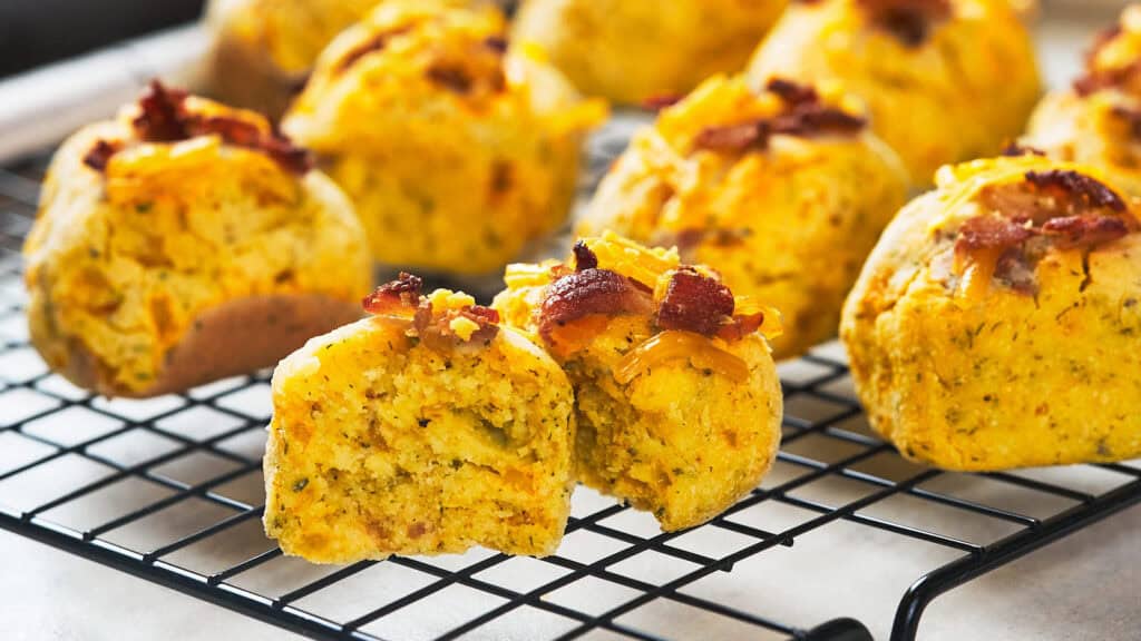 Freshly baked cheddar and bacon biscuits on a cooling rack, with one muffin cut in half to show the soft interior.