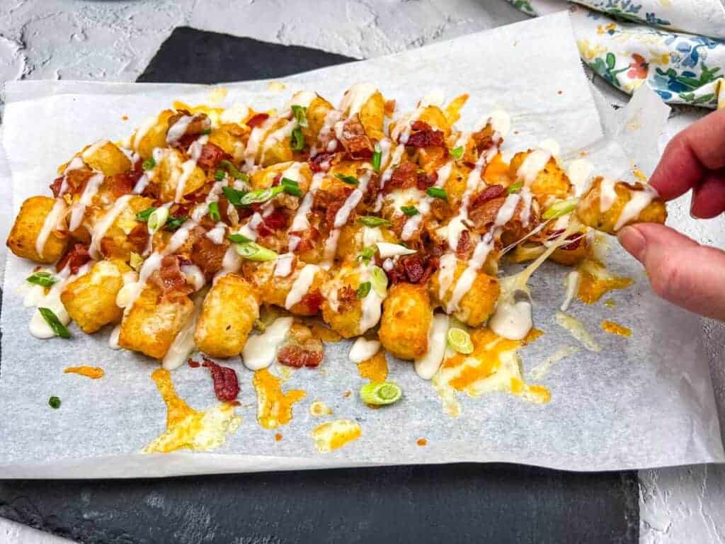 A tray of cheddar bacon loaded tater tots, drizzled with Alfredo sauce, with a hand-picking one up.