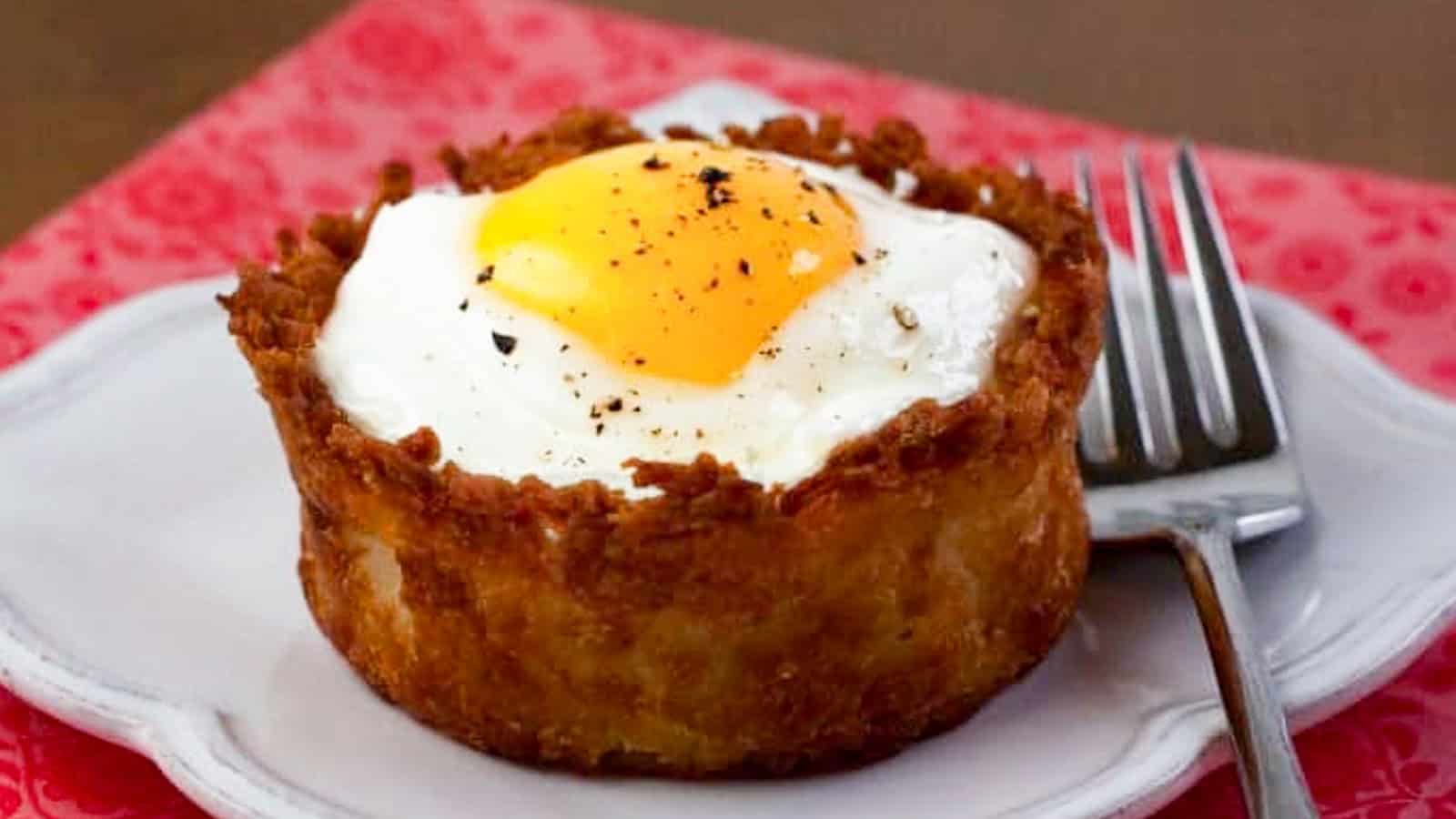A breakfast dish: crispy potato nest with a sunny-side-up egg on top, seasoned with black pepper. A fork is placed on the right side of the white plate.
