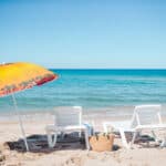 Two white lounge chairs and a straw basket under a colorful umbrella on a sunny beach with clear blue water in the background.