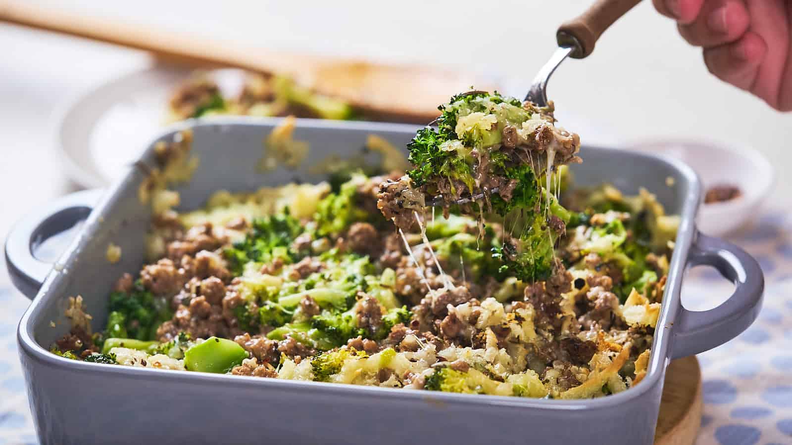 A hand lifts a portion of beef and broccoli casserole from a blue baking dish using a fork, showing melted cheese and visible broccoli and beef chunks.