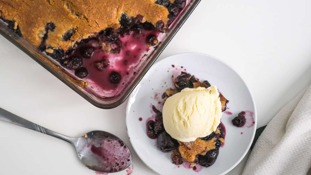 A serving of blueberry cherry cobbler with a scoop of vanilla ice cream on a white plate, next to a baking dish containing the remaining cobbler.