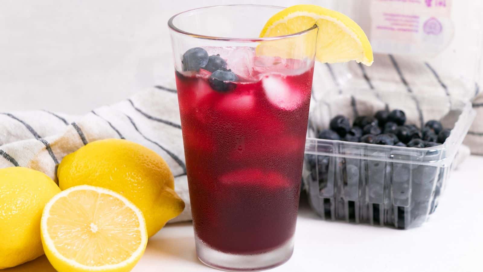 A refreshing glass of blueberry lemonade with ice, garnished with a lemon slice and blueberries, next to whole lemons and a container of blueberries.