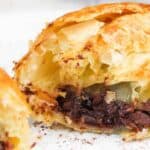 Close-up of a flaky pastry, broken open to reveal a savory minced meat filling, on a white plate.