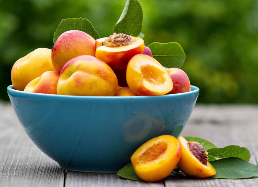 A bowl of fresh peaches, some cut in half, on a wooden table with green leaves in the background.