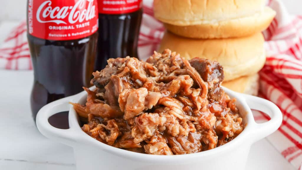 A bowl of pulled pork sits in front of two hamburger buns and two coca-cola bottles on a checkered red and white cloth.