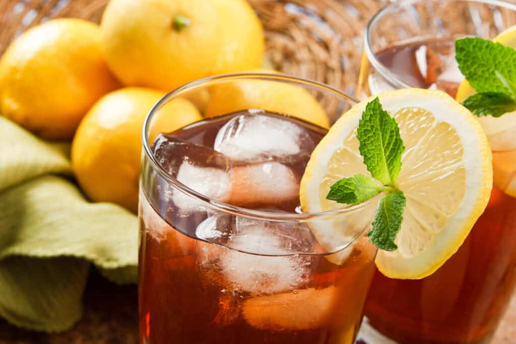 Two glasses of sweet tea with ice cubes, garnished with lemon slices and mint leaves, are placed in front of a basket of lemons.