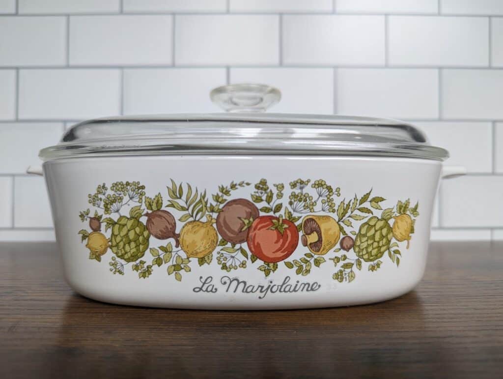 White ceramic casserole dish with a glass lid, adorned with illustrations of fruits and herbs. The vintage CorningWare piece is labeled "La Marjolaine" and rests on a wooden counter against a subway tile backsplash.